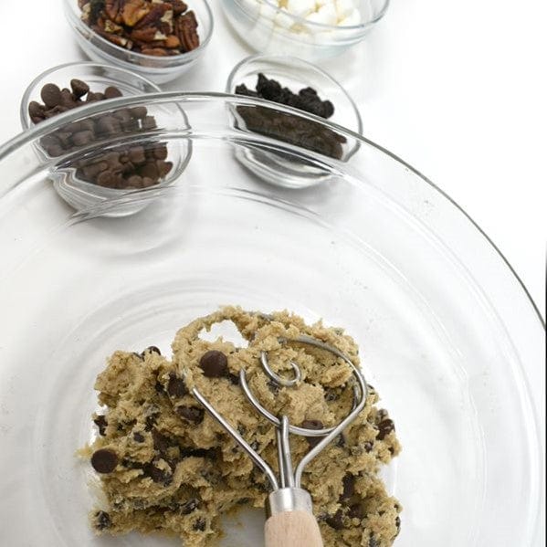 Norpro 12'' Danish Dough Whisk being used to mix up chocolate chip cookie dough in clear glass mixing bowl with add-ins like chocolate chips, nuts, marshmallows, and raisins sitting in glass bowls behind cookie dough bowl