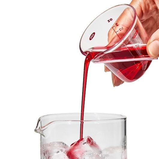 small measuring cup with milliliter markings pouring red liquid into glass with ice