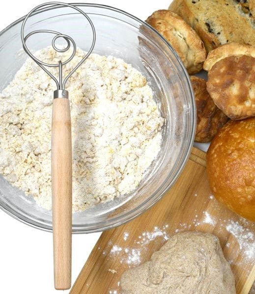 closeup of Norpro 12'' Danish Dough Whisk sitting on top of clear glass mixing bowl with dry ingredients inside and sourdough bread baked goods behind it with bread board and unbaked loaf of sourdough on top