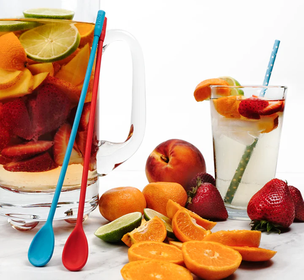  side view of two long handle silicone spoons leaning on pitcher next to peaches, oranges, limes, and strawberries, with pitcher of fruit juice and glass with juice and straw