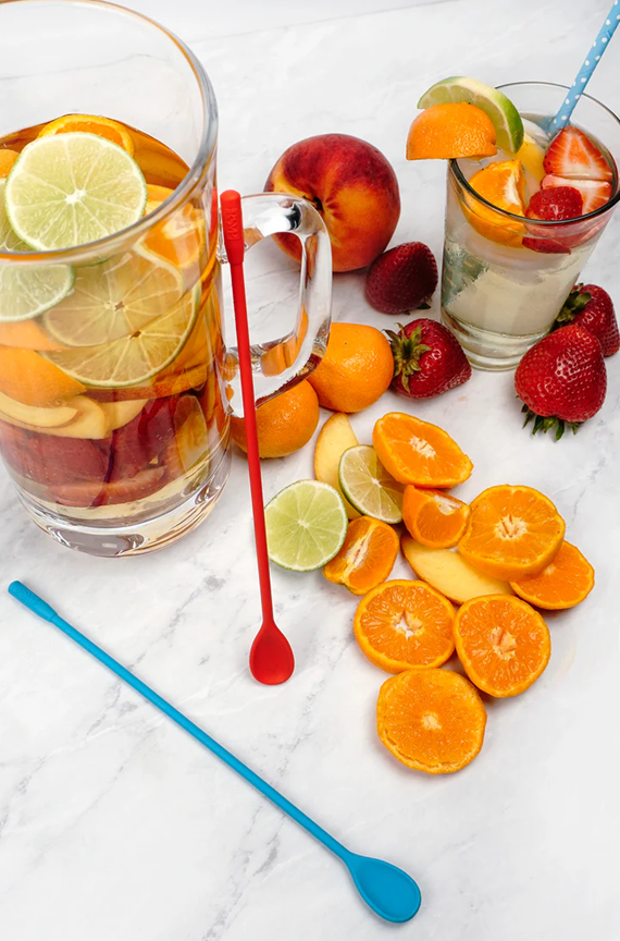 top view of two long handle silicone spoons leaning on pitcher next to peaches, oranges, limes, and strawberries, with pitcher of fruit juice and glass with juice and straw