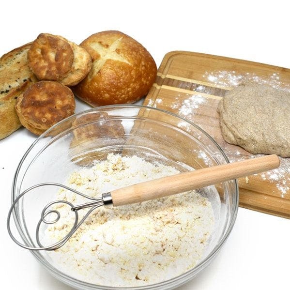 Norpro 12'' Danish Dough Whisk sitting on top of clear glass mixing bowl with dry ingredients inside and sourdough bread baked goods behind it with bread board and unbaked loaf of sourdough on top