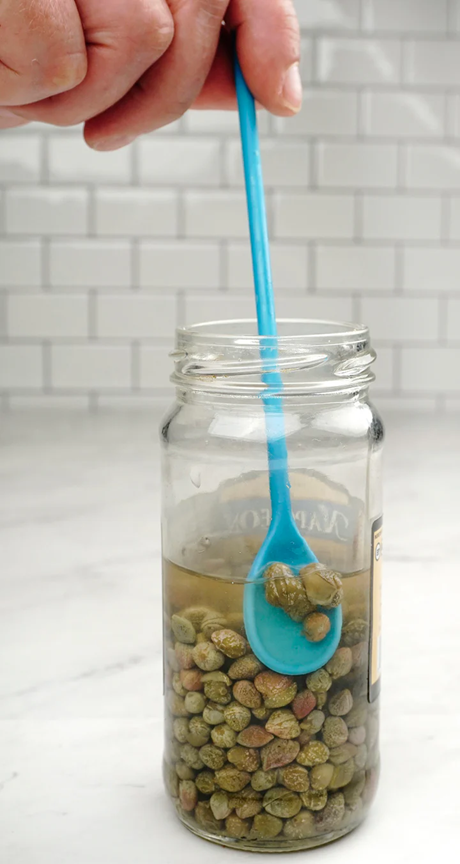 closeup of blue long handled spoon scooping capers out of jar on white kitchen counter