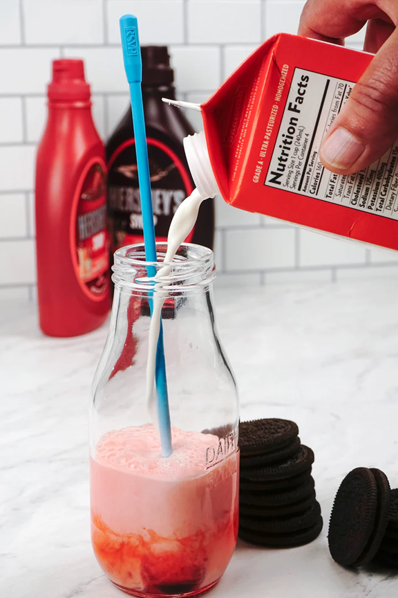 blue long handle stirrer spoon mixing a strawberry milkshake on kitchen counter with milk pouring into jar next to chocolate cookies