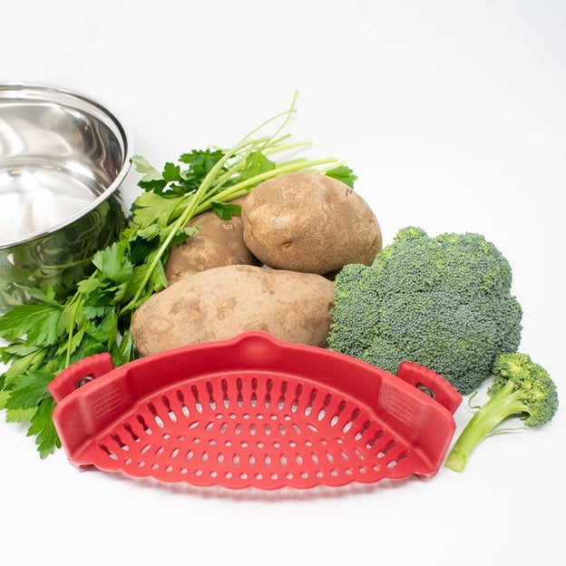 red silicone vegetable strainer in front of silver pot, herbs, and raw potatoes and broccoli